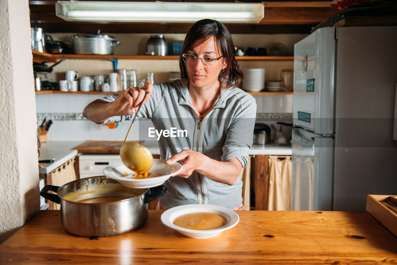 Housewife pouring delicious aromatic pumpkin cream soup into plate while serving dinner in home kitchen