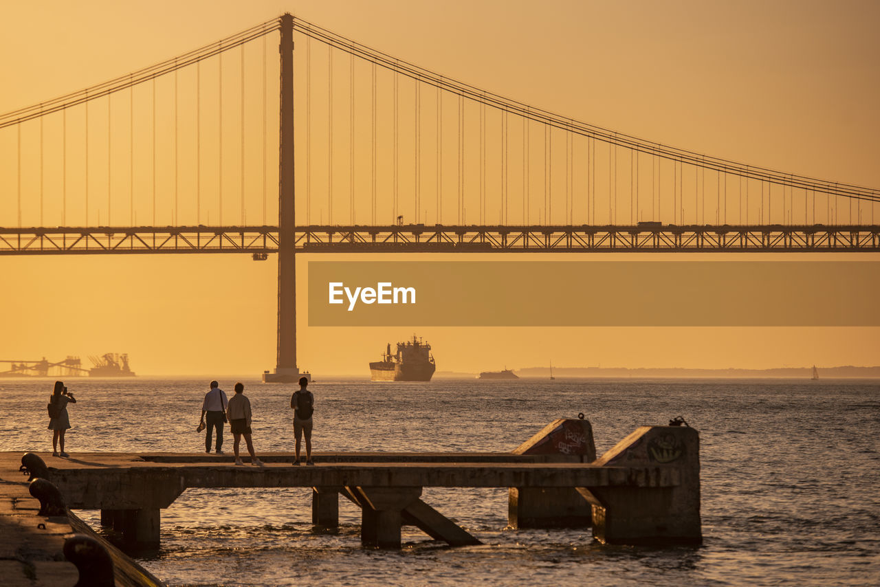 rear view of people walking on suspension bridge against clear sky during sunset