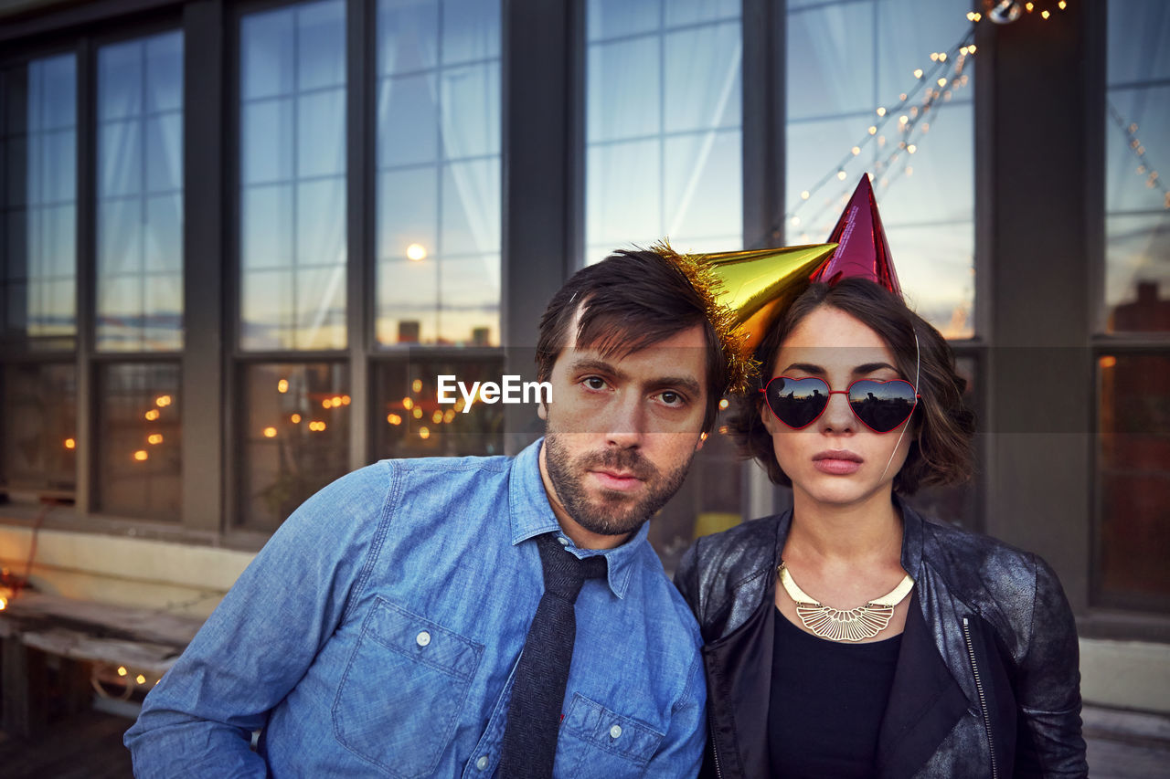 Portrait of friends wearing party hats standing on building terrace against glass windows