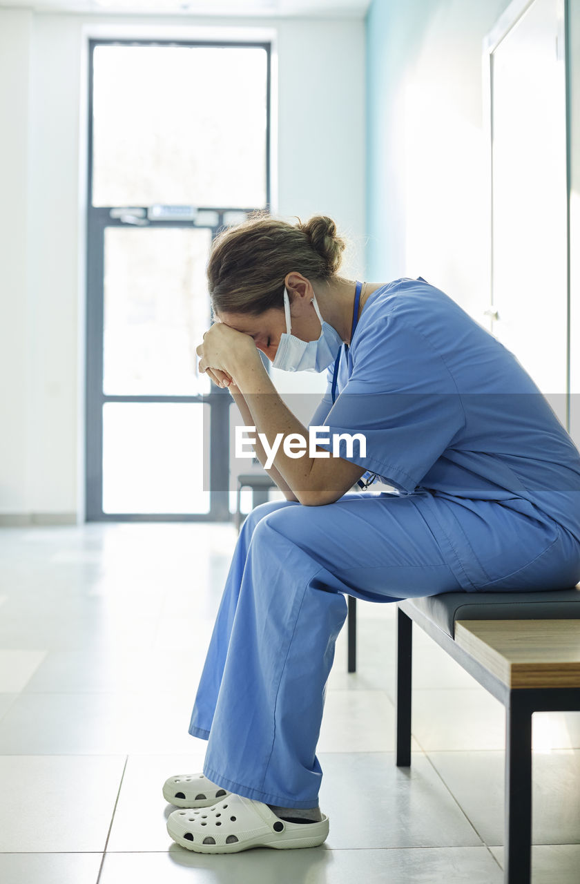 Exhausted nurse wearing protective face mask sitting on bench in corridor