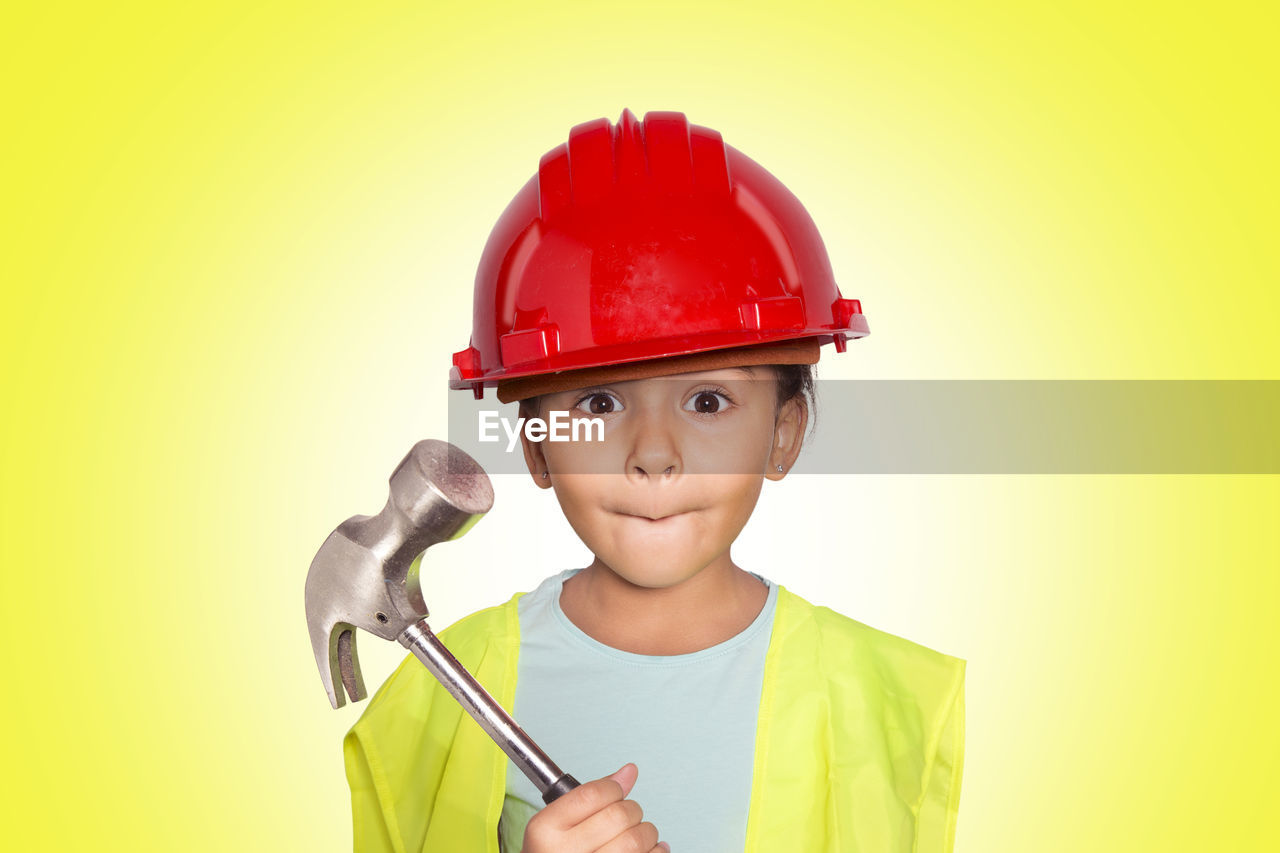 Portrait of cute girl holding hammer while wearing helmet against yellow background