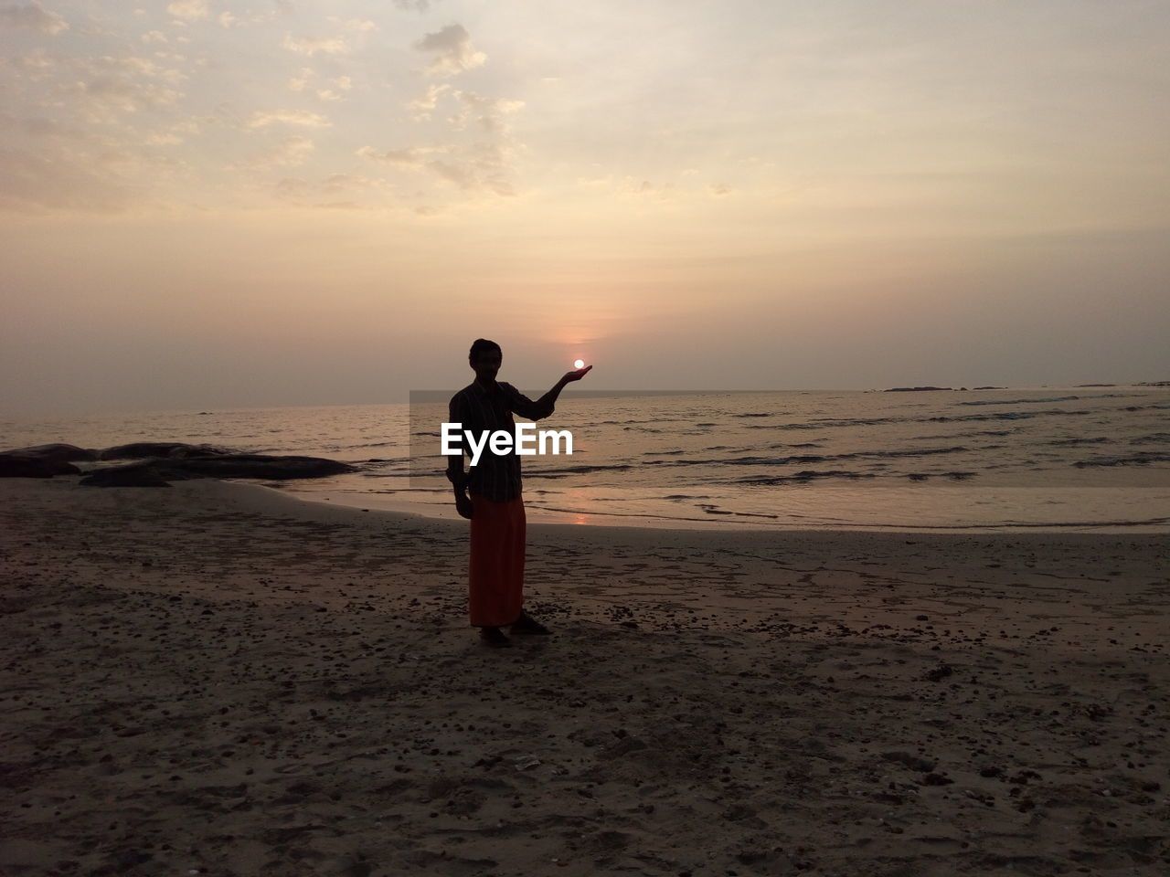 Optical illusion of silhouette man holding sun while standing at beach against sky