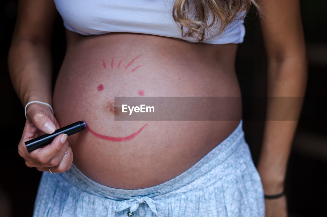 Midsection of pregnant woman drawing on stomach against black background