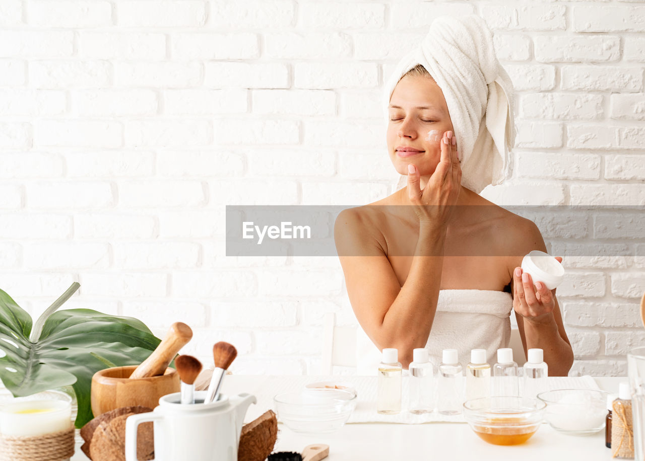  happy young woman wearing white bathrobes towels on head doing spa procedures applying creme