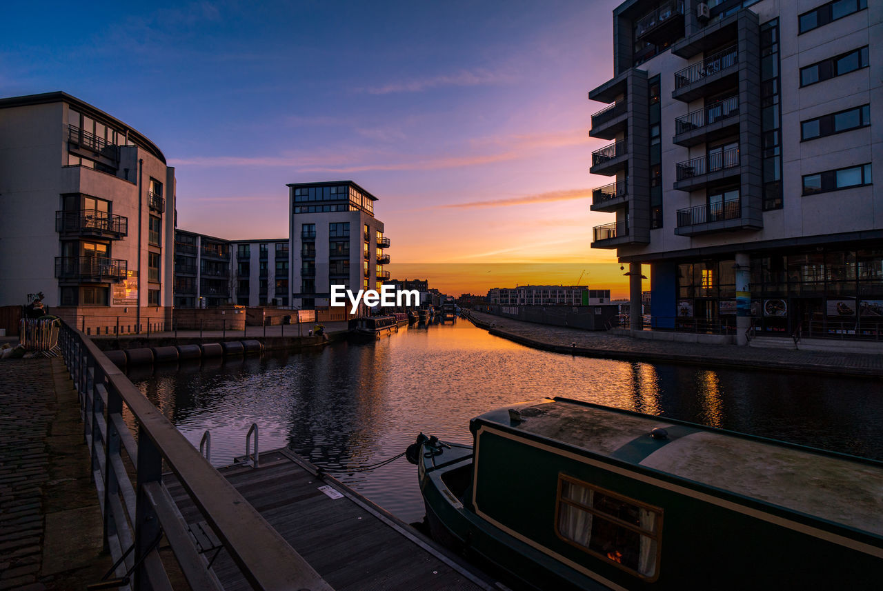 BUILDINGS BY RIVER AGAINST SKY AT SUNSET