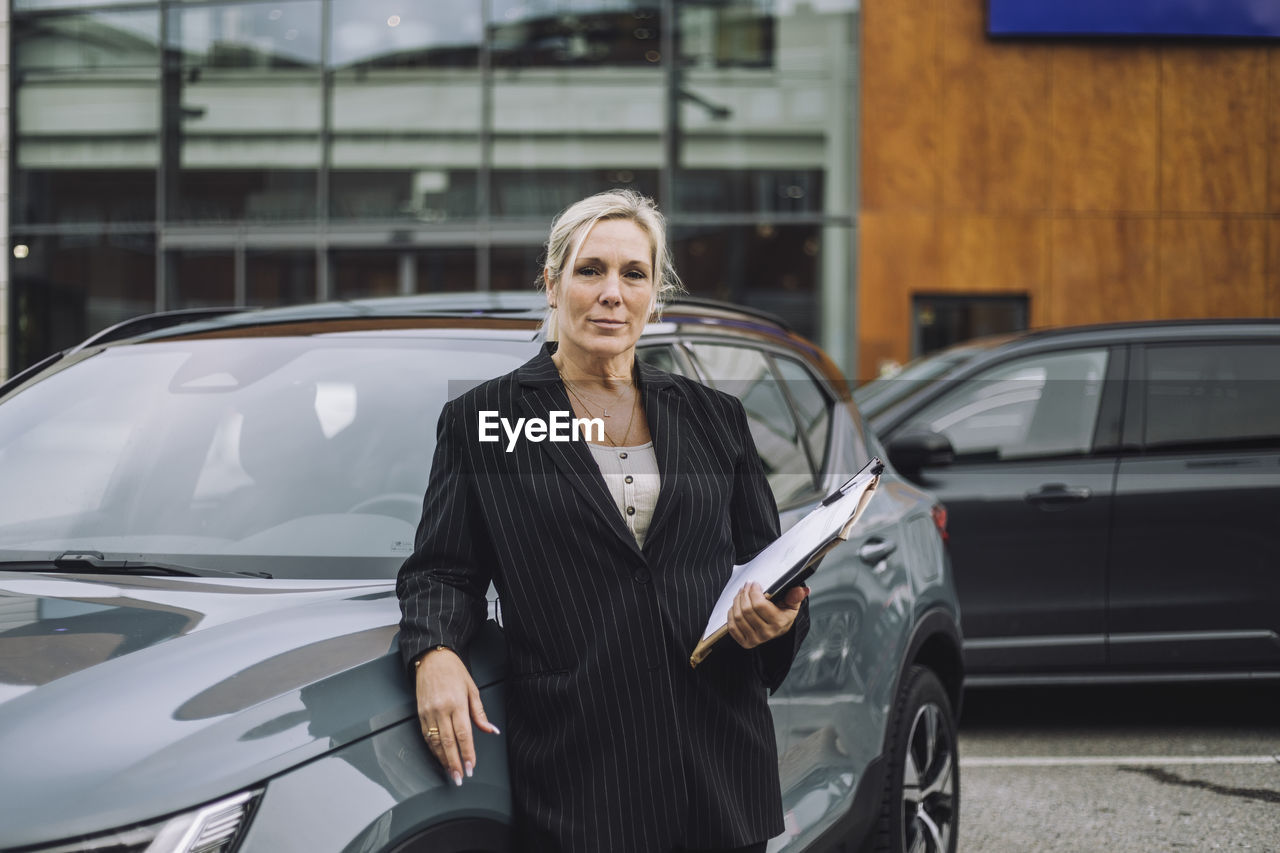 Portrait of confident saleswoman with documents standing next to car