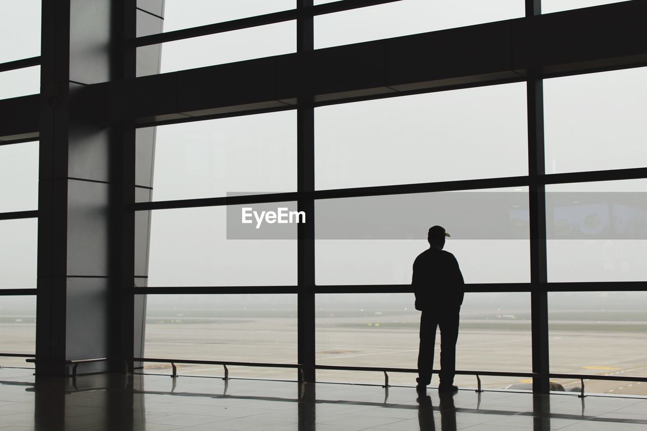 Rear view of silhouette man looking through window while standing at airport