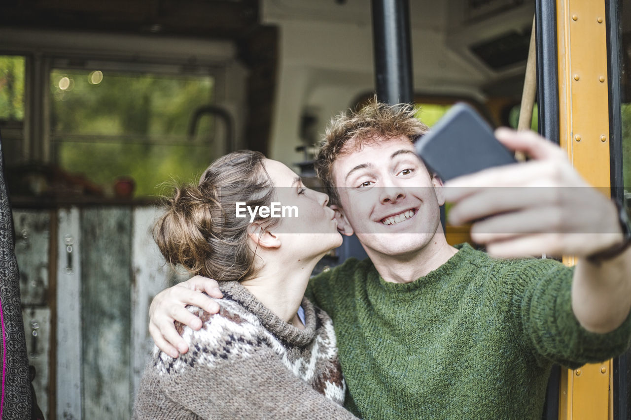 Smiling man taking selfie from mobile phone while woman kissing him during camping