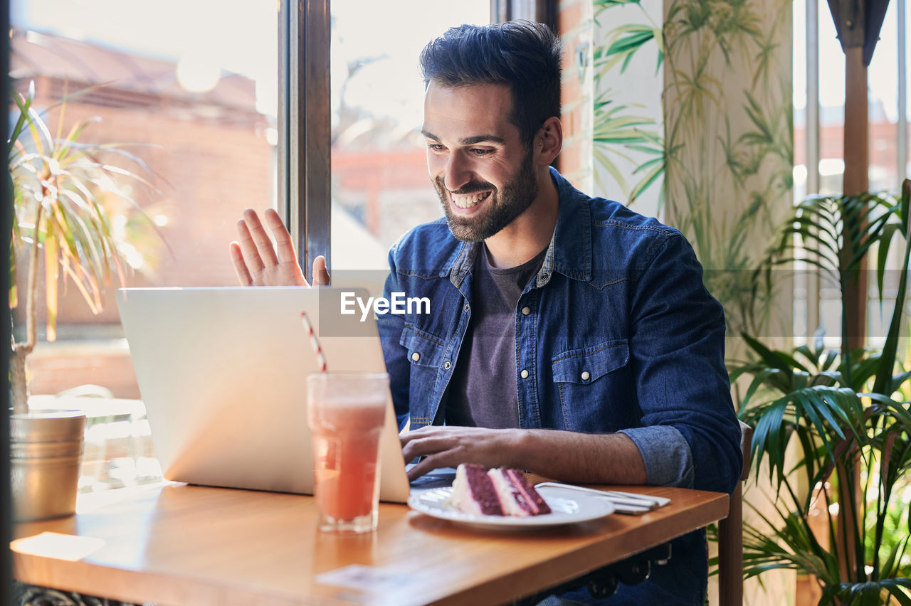 Cheerful ethnic male waving hand during video chat on netbook at table with tasty cake piece and refreshing drink