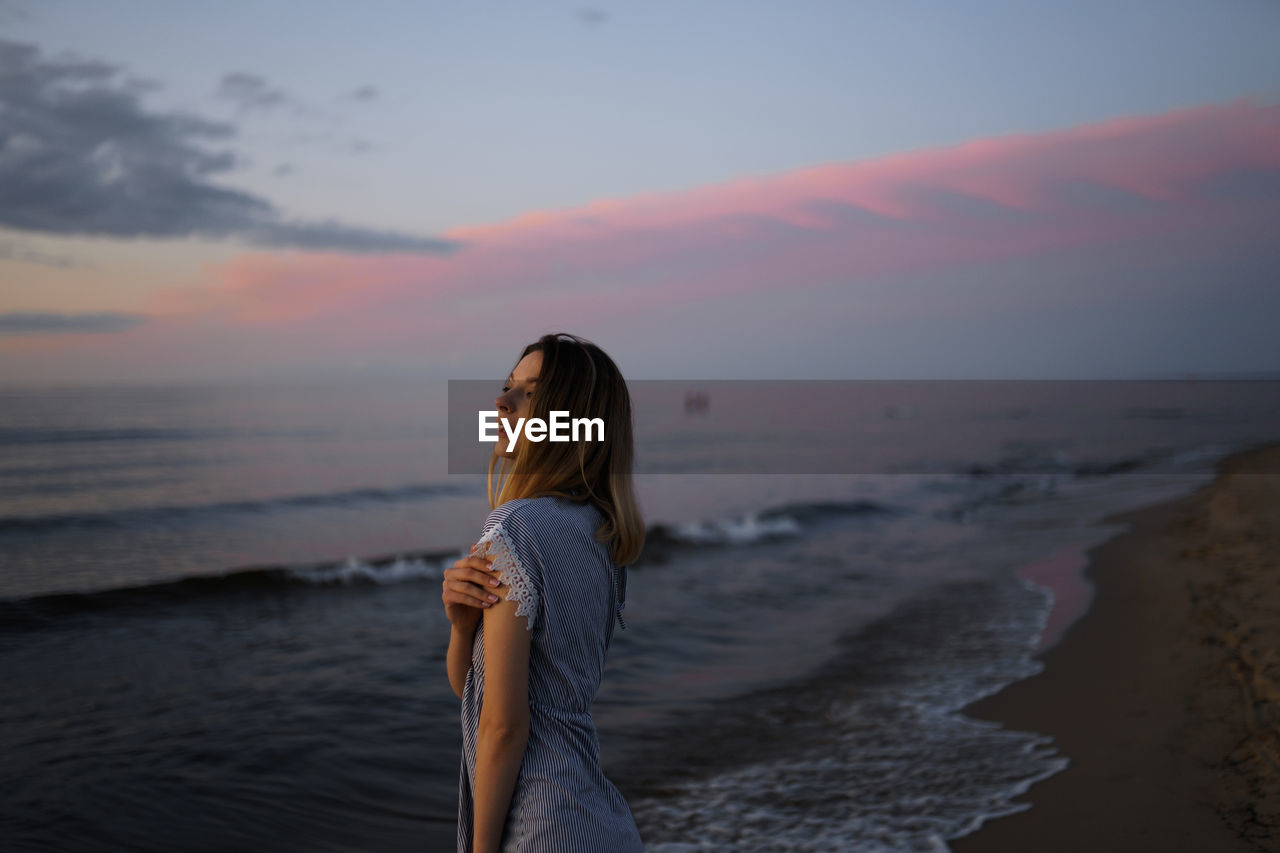 Woman looking away while standing at beach during sunset