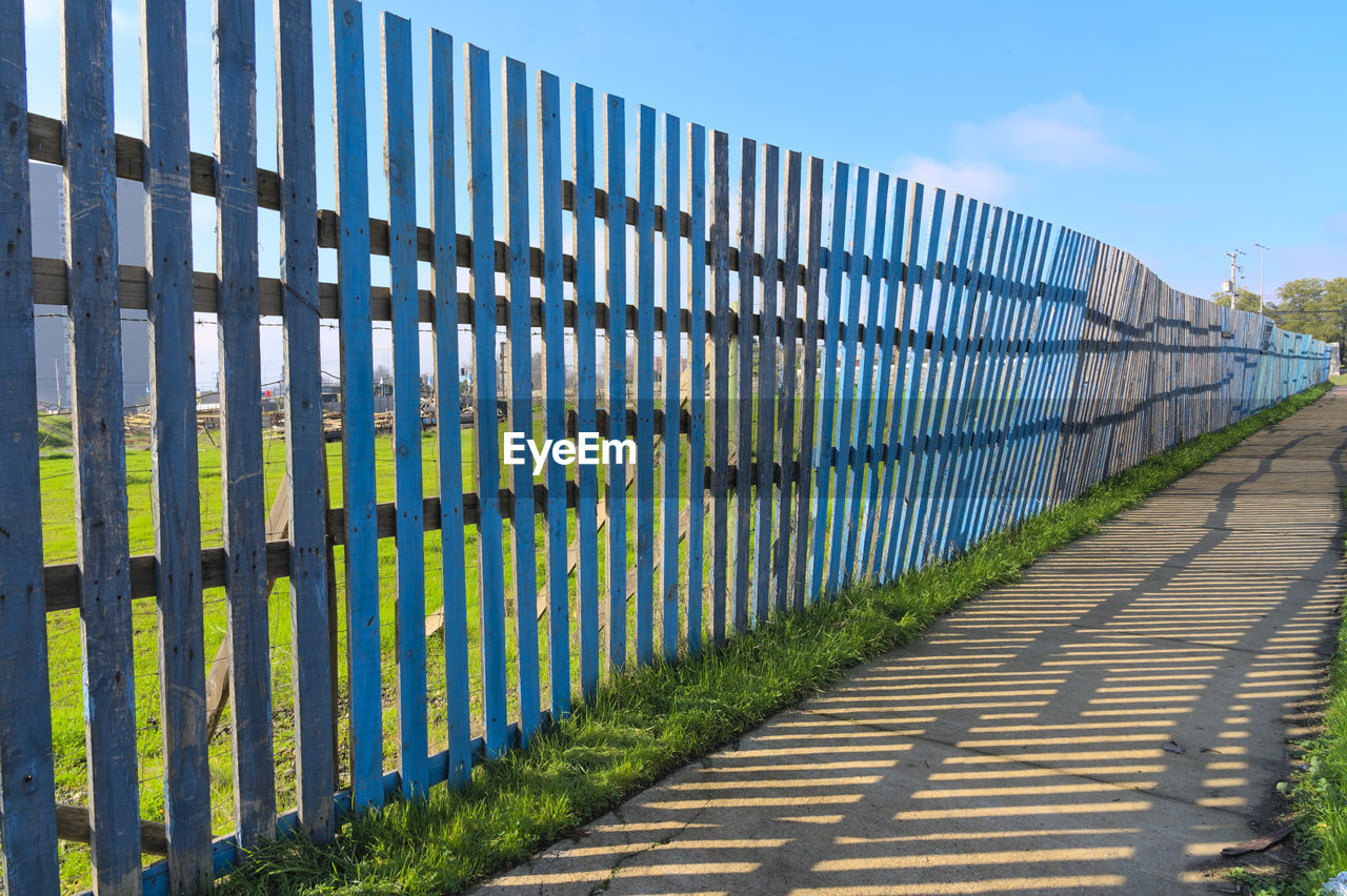 fence, home fencing, outdoor structure, security, protection, sky, nature, no people, day, architecture, blue, plant, footpath, outdoors, picket fence, sunlight, walkway, in a row, wall, cloud, grass, diminishing perspective, land, shadow, wood, built structure, gate