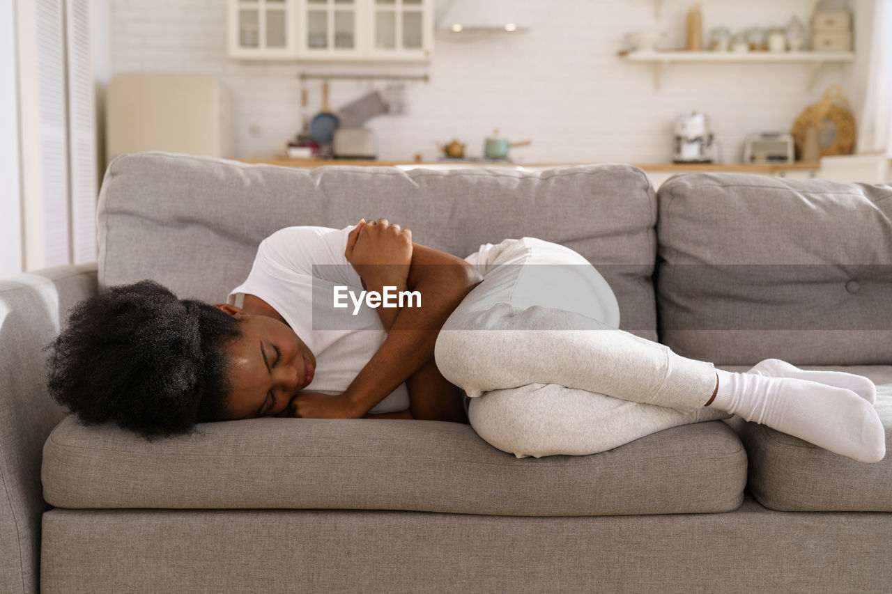 Depressed black girl with crossed arms lying on couch at home crying, suffering from breakup.