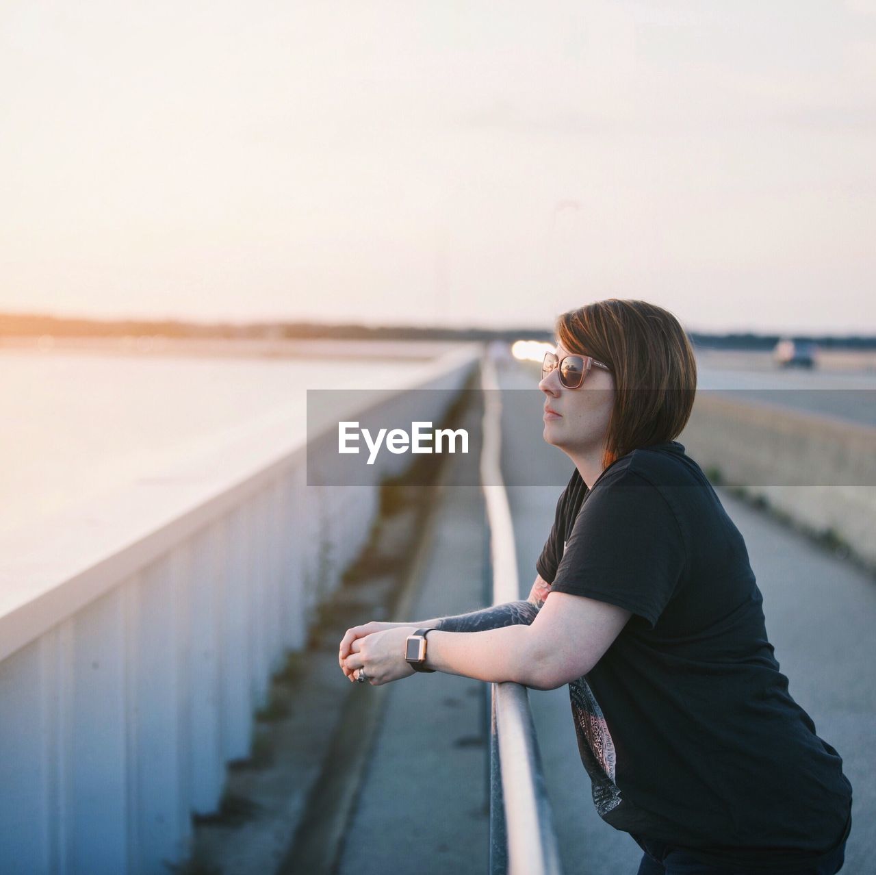 Side view of young woman wearing sunglasses standing by railing against sky during sunset