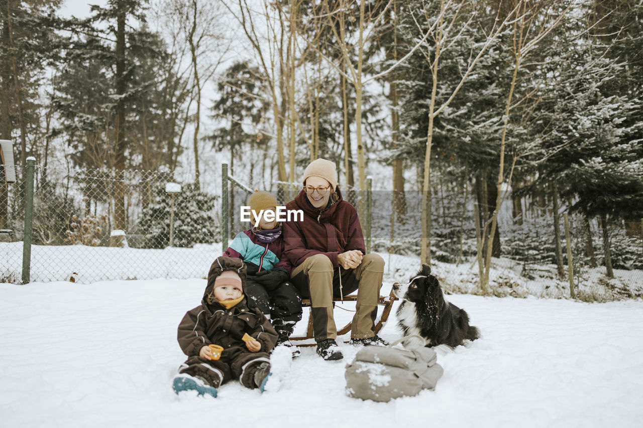 Mother with daughters and border collie sitting on snow covered field against trees