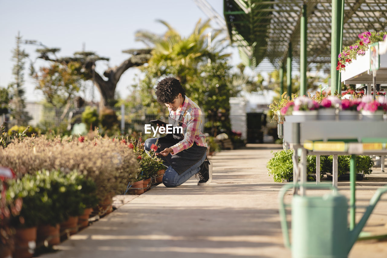 Florist with tablet pc kneeling near plants at nursery