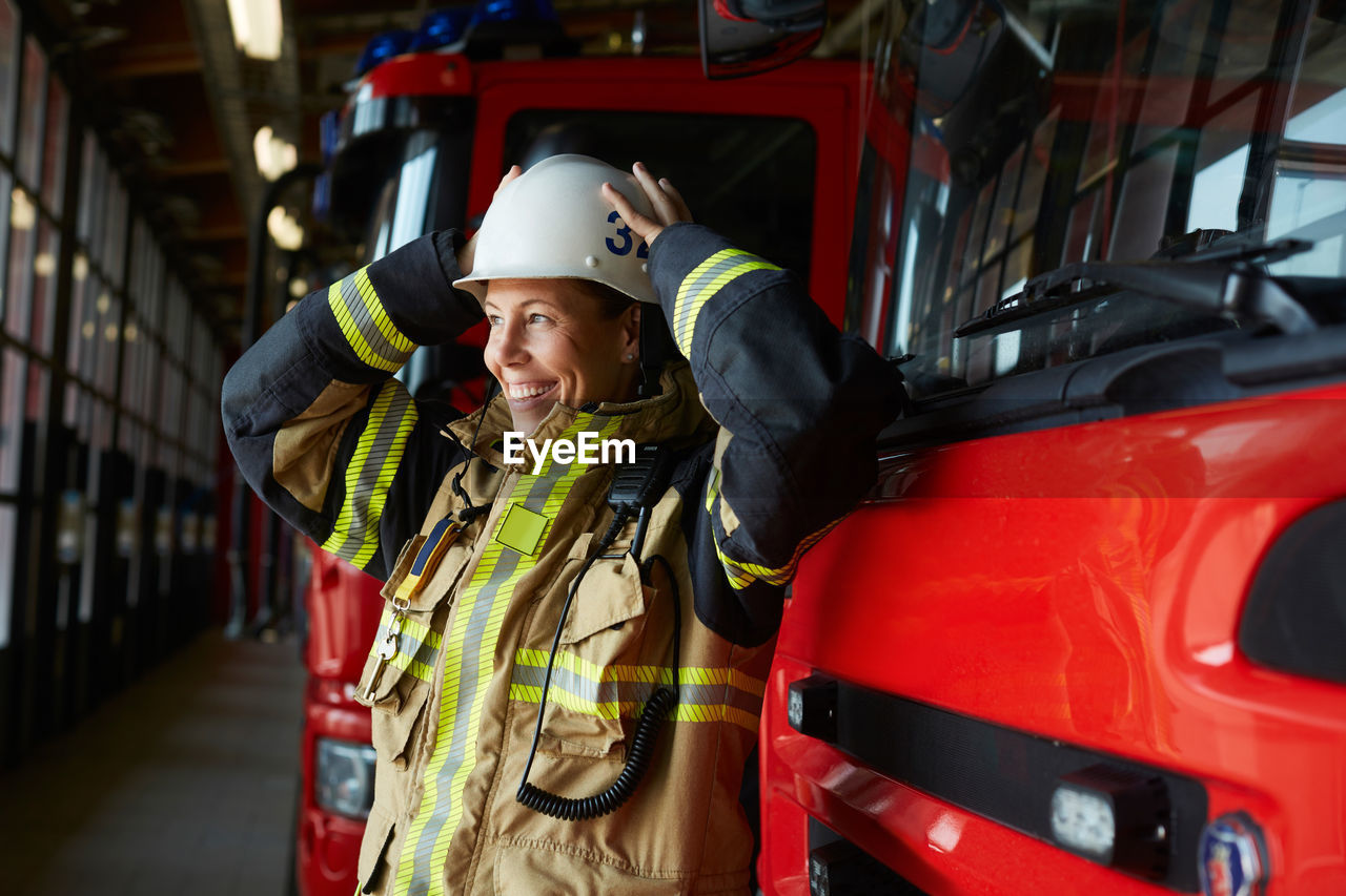 Smiling female firefighter wearing helmet while standing by fire engine at fire station