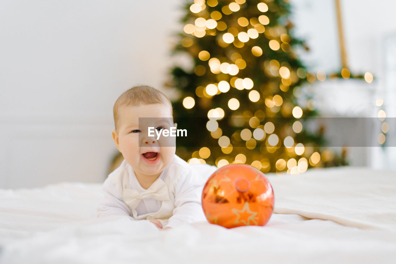 The baby laughs, covering one eye, lying on the bed against the background of christmas lights