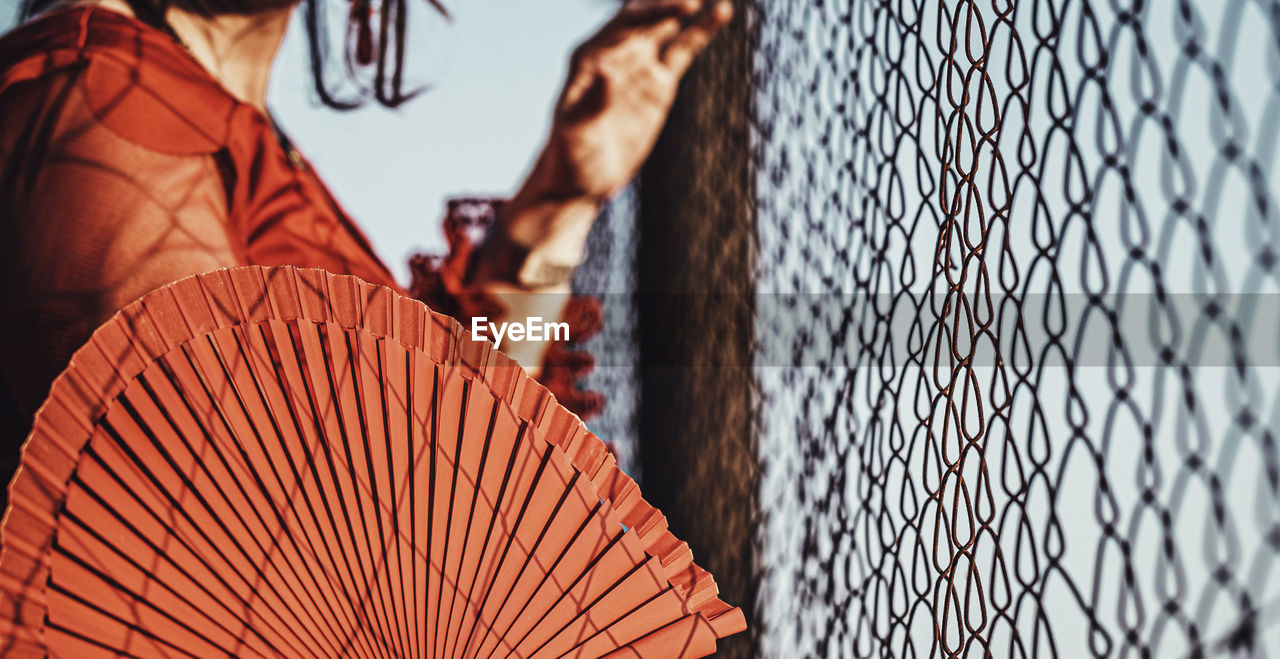 Midsection of woman holding hand fan by chainlink fence