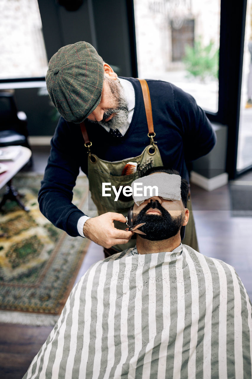 Concentrated middle aged unshaven male barber in apron and beret cutting dark beard of ethnic client with scissors in traditional barbershop