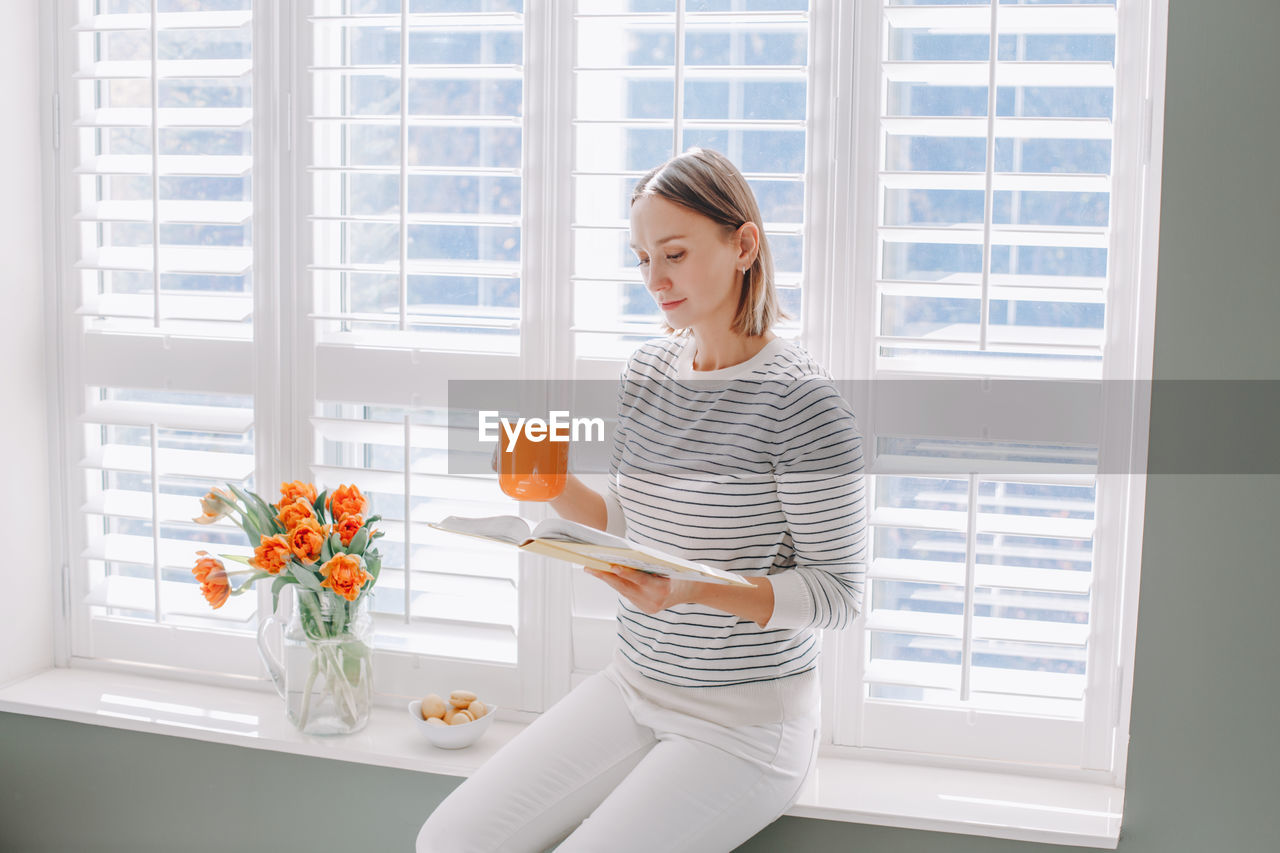 Woman reading book while sitting by window at home