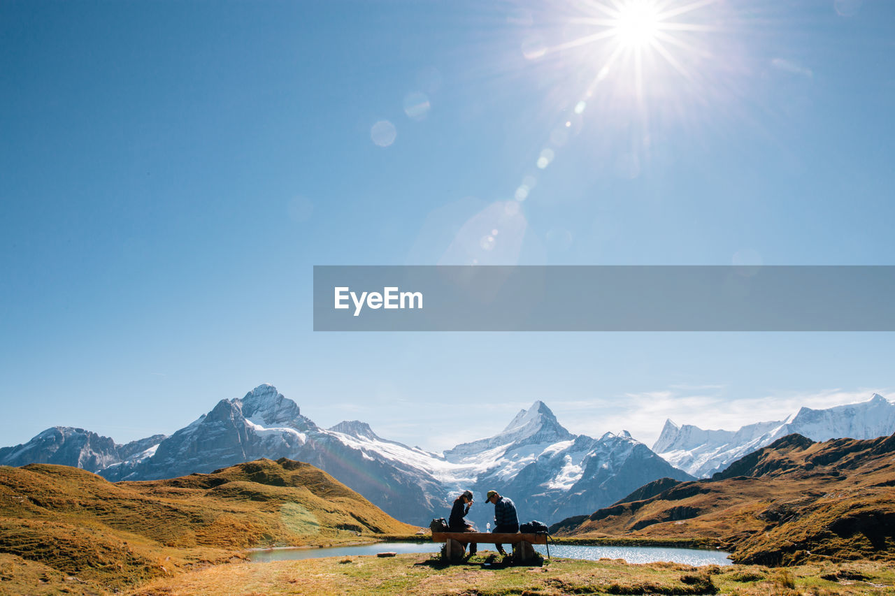 People sitting on bench by snowcapped mountains against sky