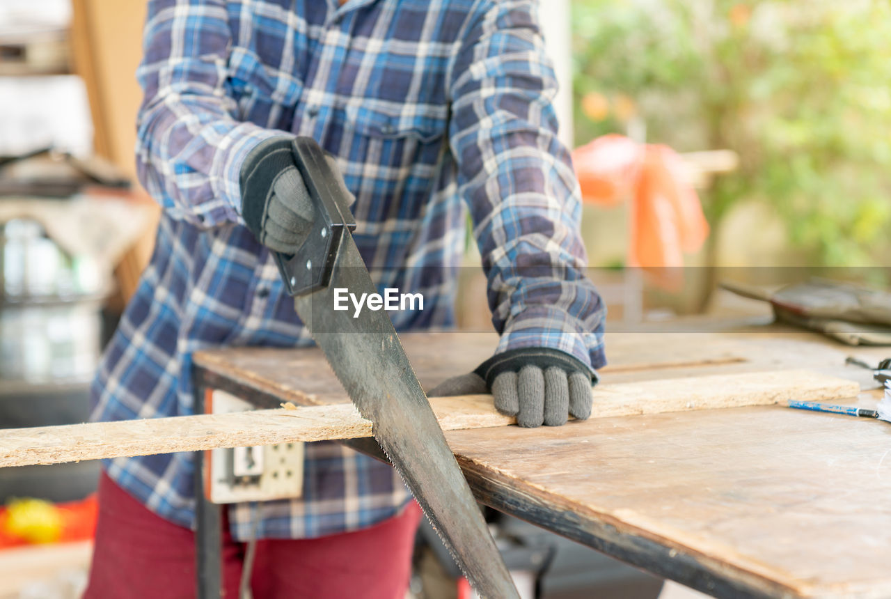 A carpenter ware gloves use hand saw to sawing wood on a table in a workshop.