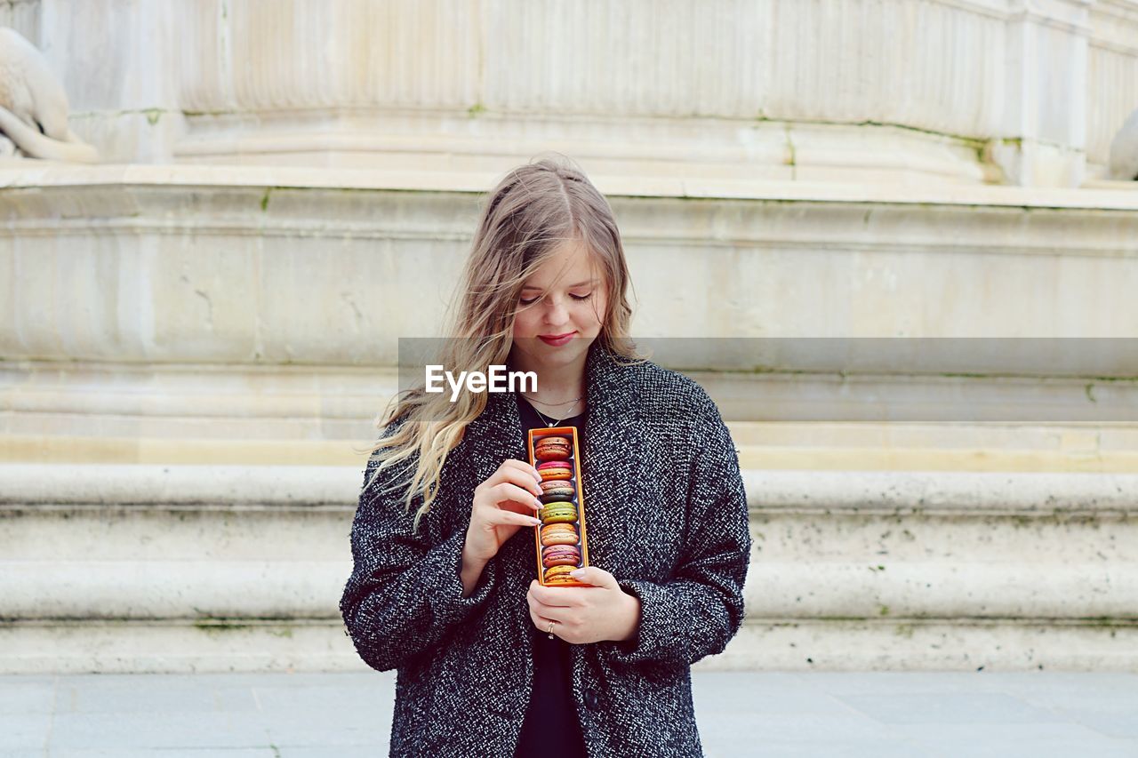 Young woman with macaroons standing against wall