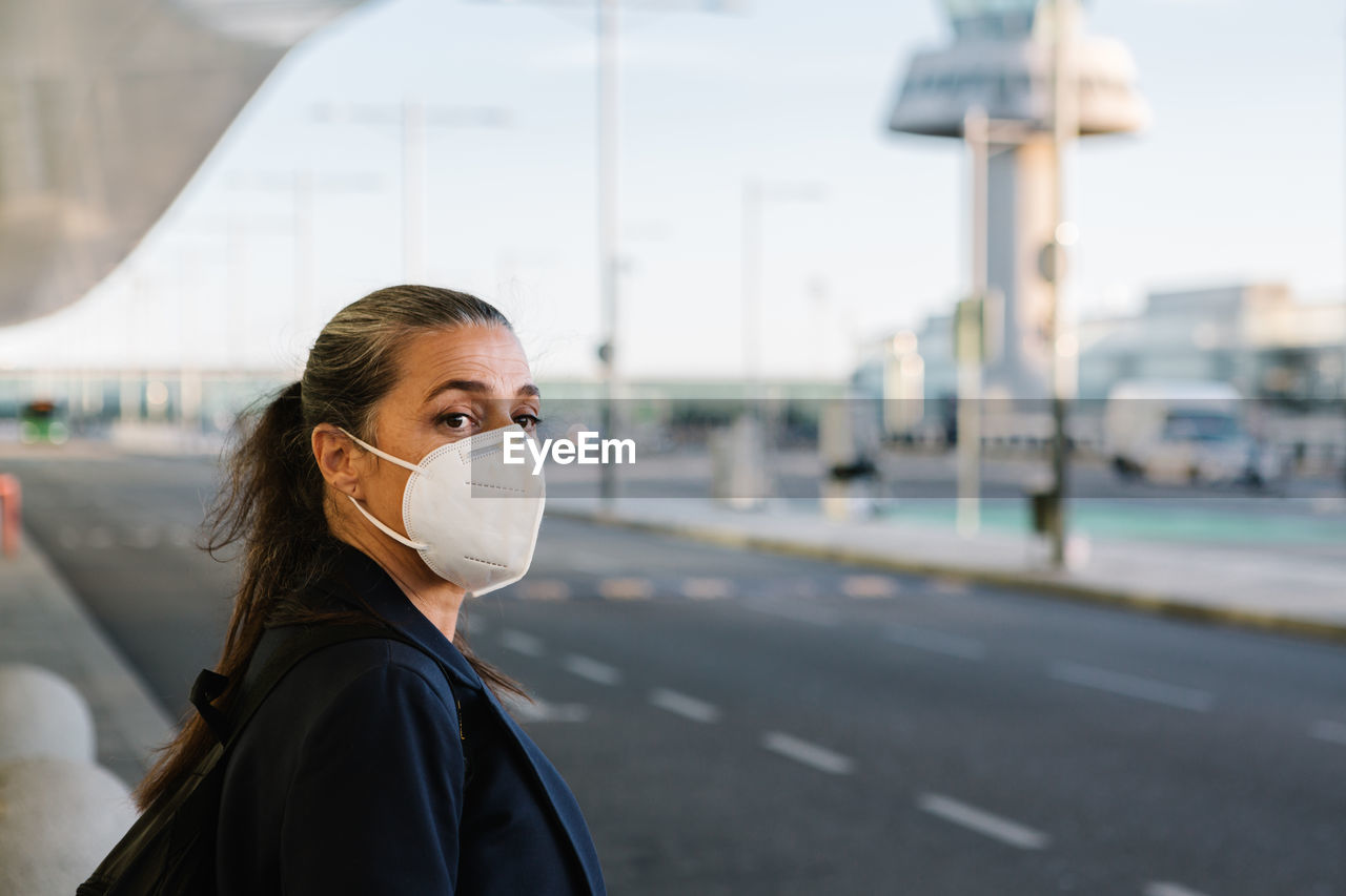 Female tourist in protective respirator during covid 19 epidemic standing in airport and catching taxi with outstretched arm while looking away
