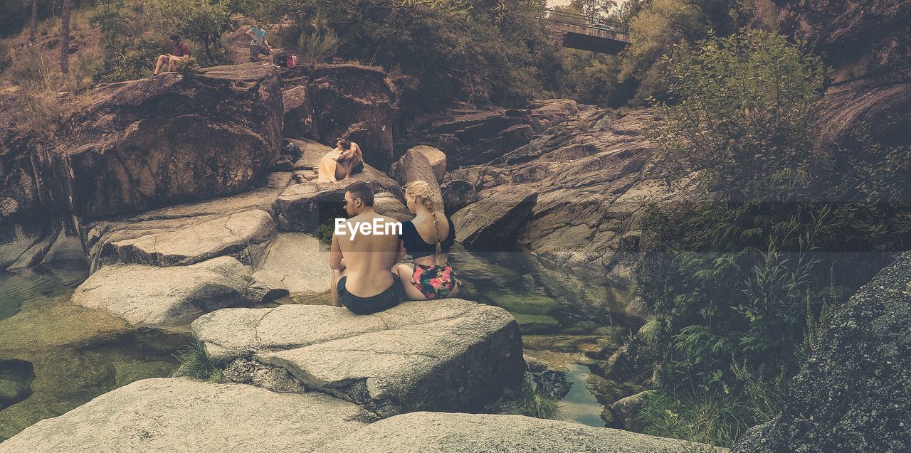 REAR VIEW OF FRIENDS SITTING ON ROCK