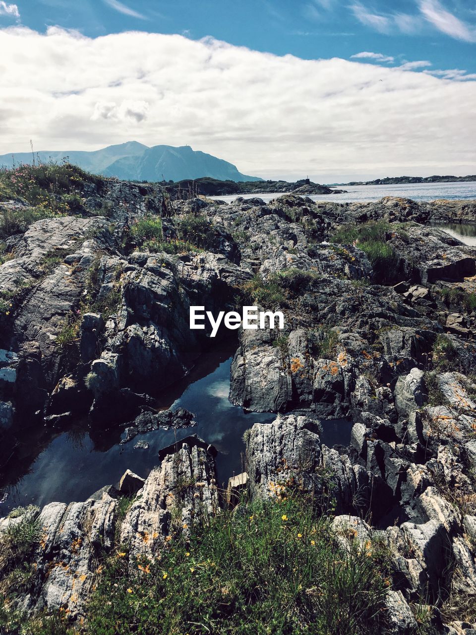 Scenic view of sea and rocky mountains against cloudy sky during sunny day