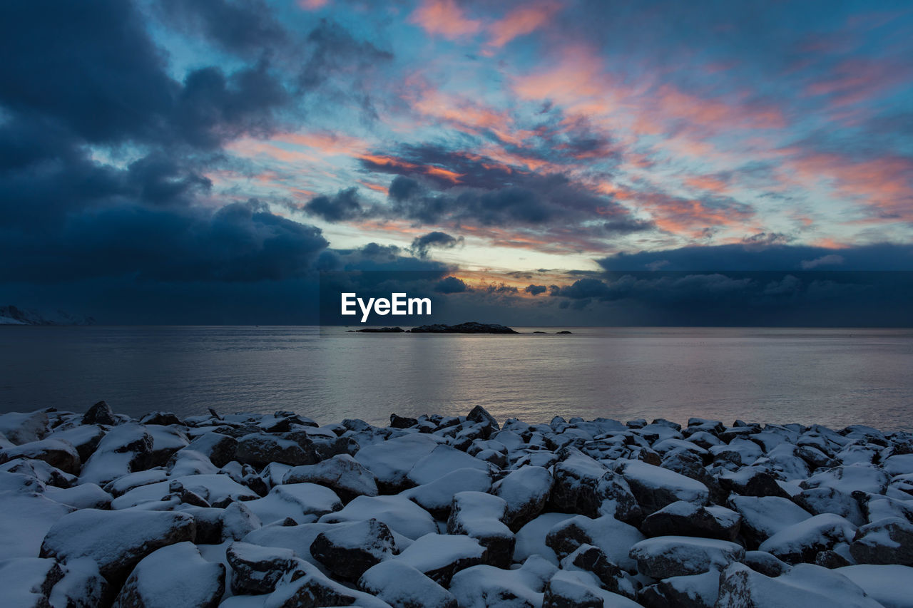 Snow covered rocky shore by sea against cloudy sky during sunset