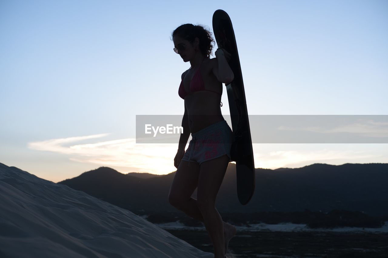 Beautiful woman walking with sandboard at her back at sunset