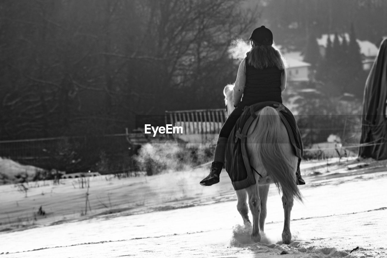 Full length rear view of female jockey riding horse on snow covered field