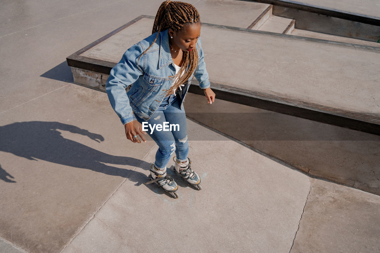 From above of cool african american female in denim clothes rollerblading in skate park on sunny day in summer