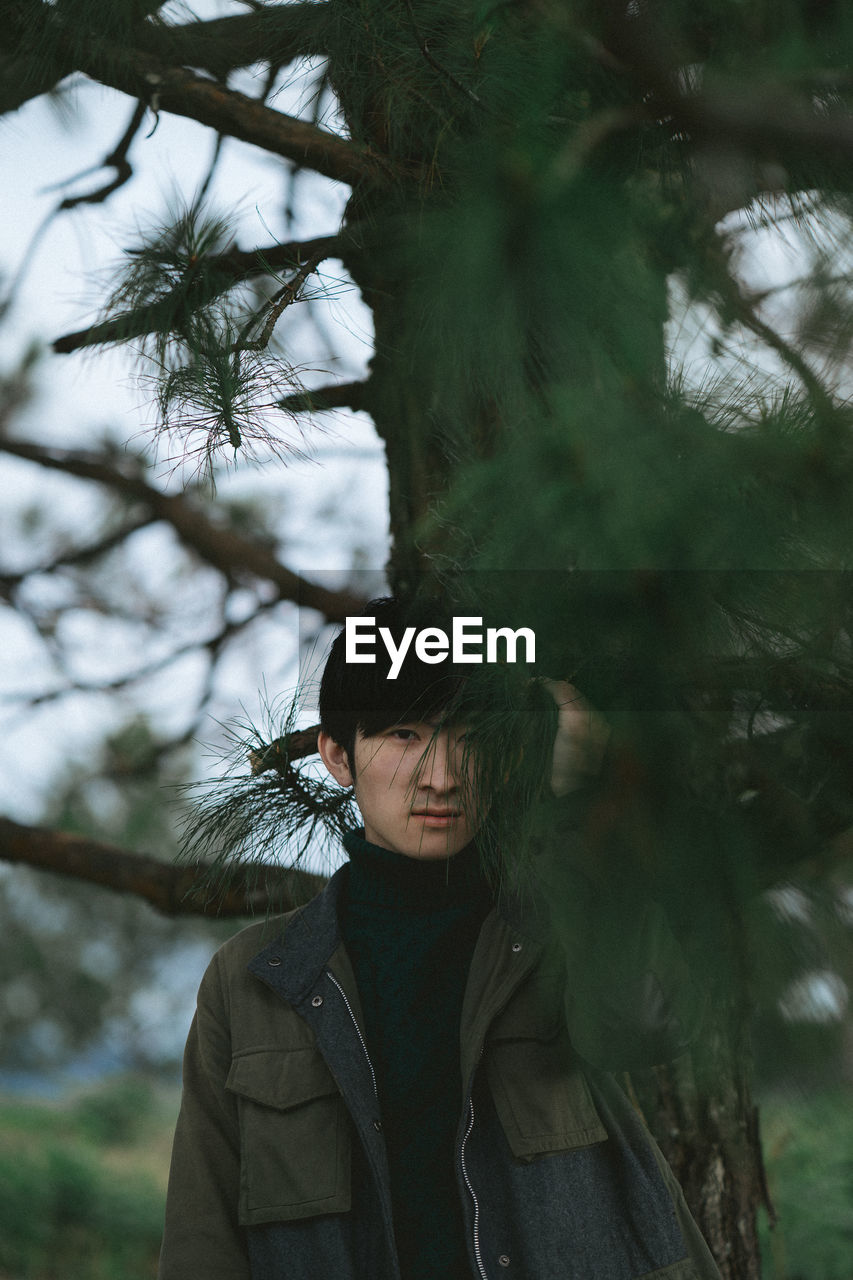 Portrait of man seen through leaves standing against tree trunk