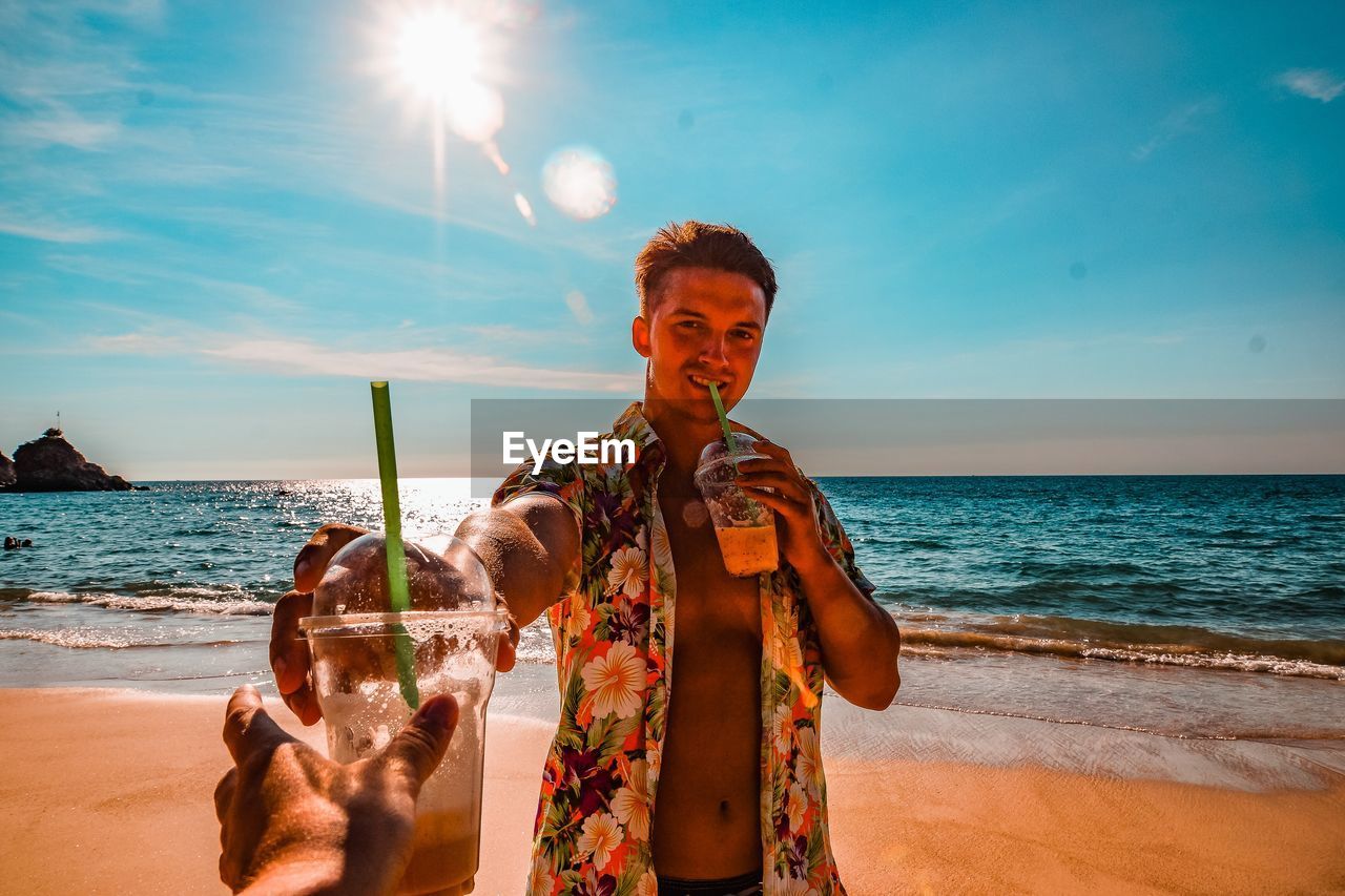 Cropped hand of person receiving drink from man drinking juice at beach