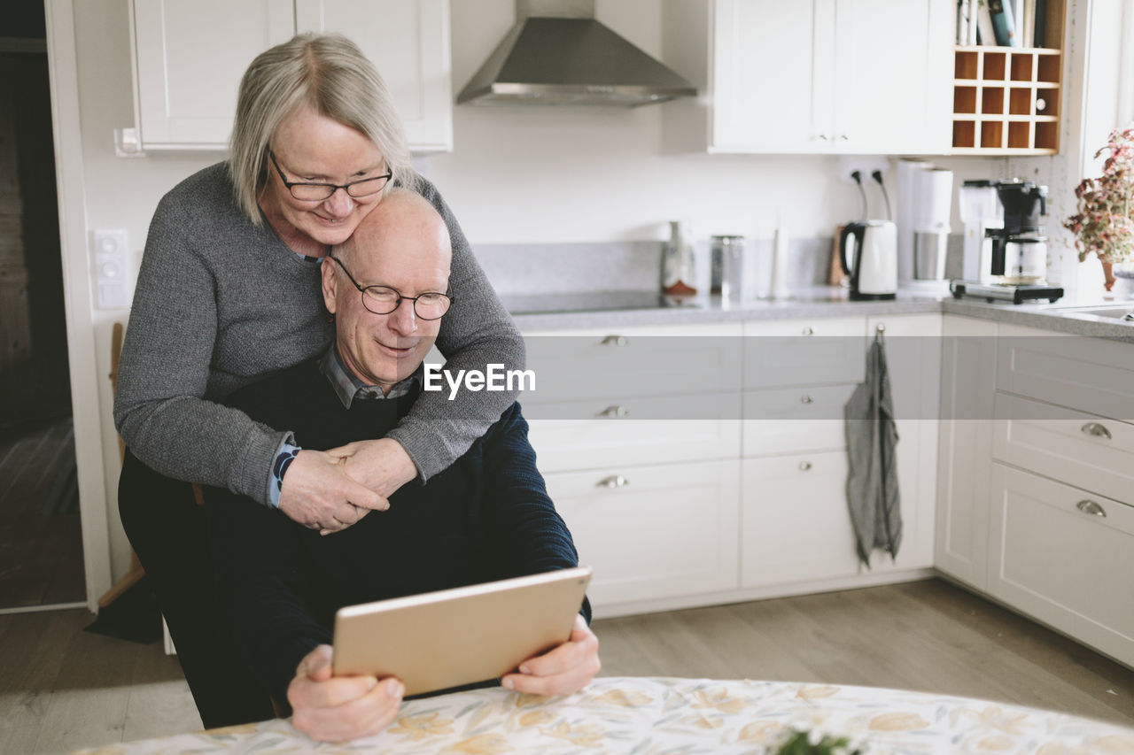 Smiling couple embracing and using tablet at kitchen table