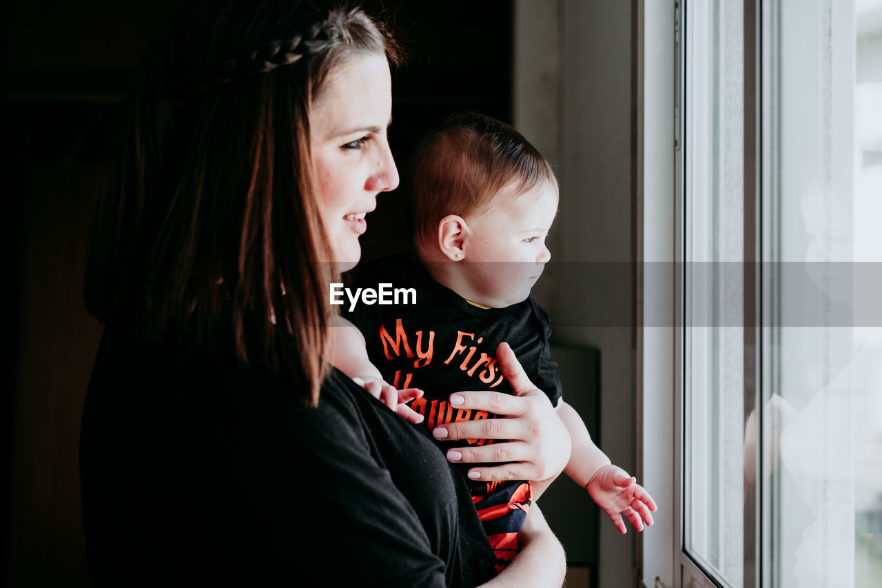 Mother and daughter embracing while standing by window at home