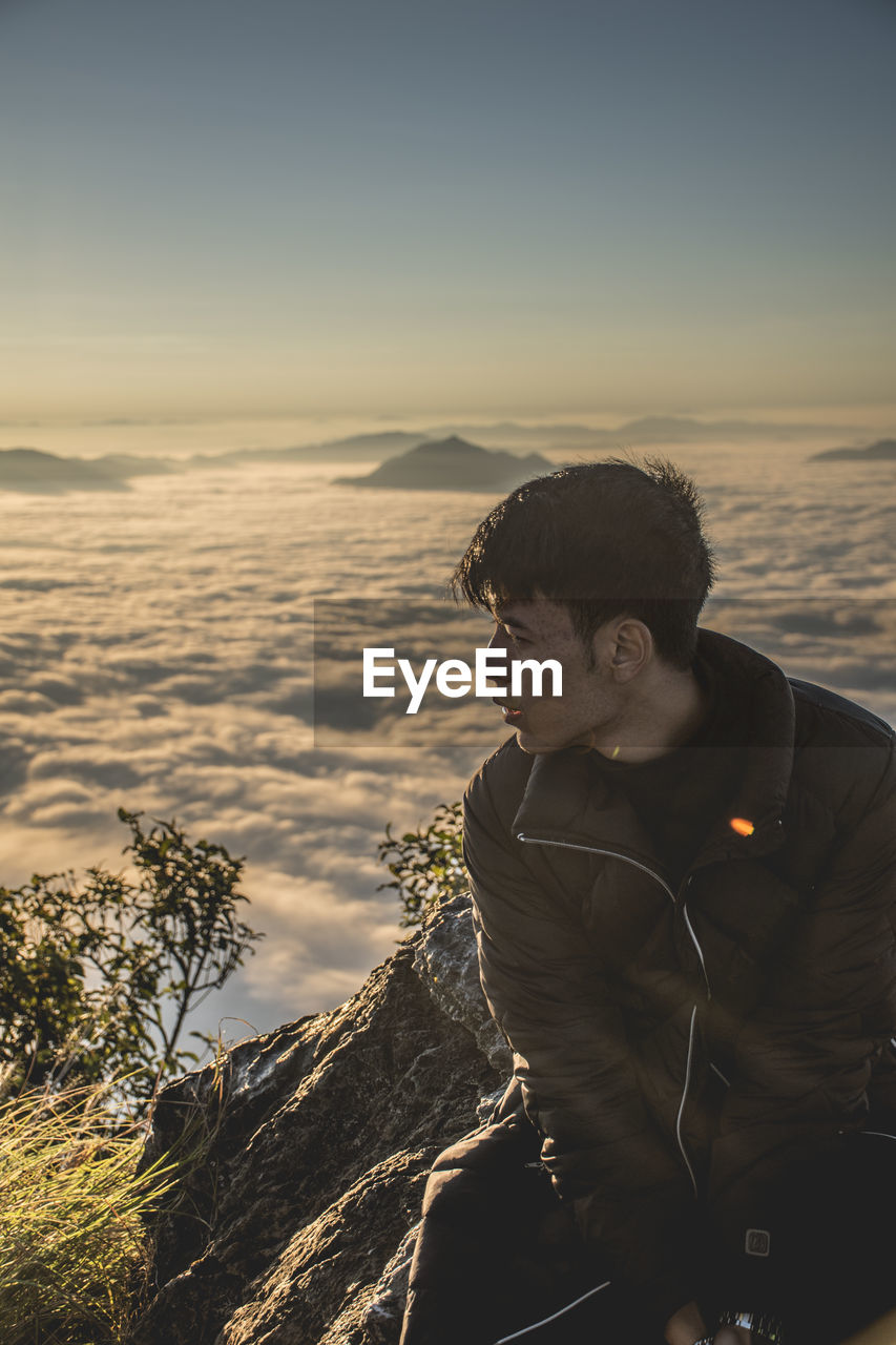 Young man looking away while sitting on mountain against clear sky during sunset