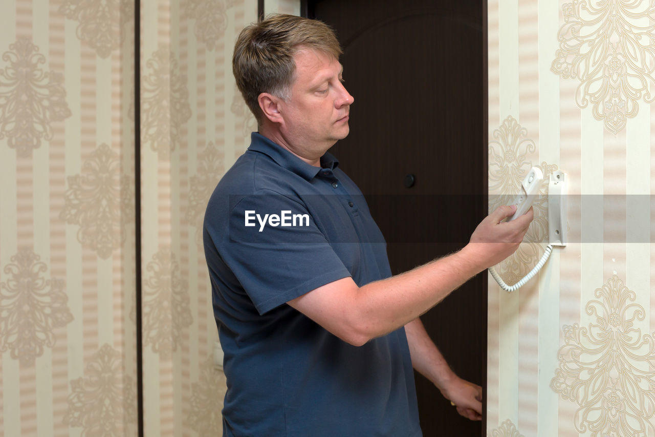 An adult blond man is talking intercom in the hallway of his apartment.