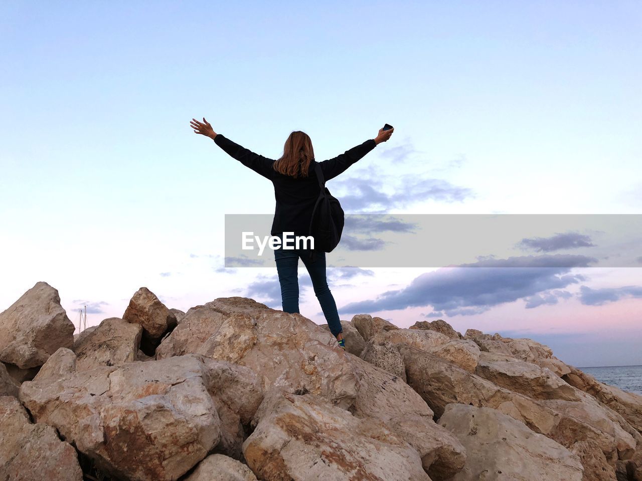 Rear view of woman with arms outstretched standing on rocks against sea during sunset