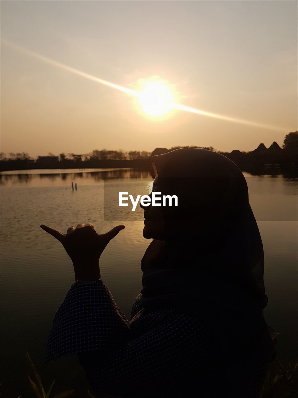 Woman gesturing shaka sign by lake against sky during sunset