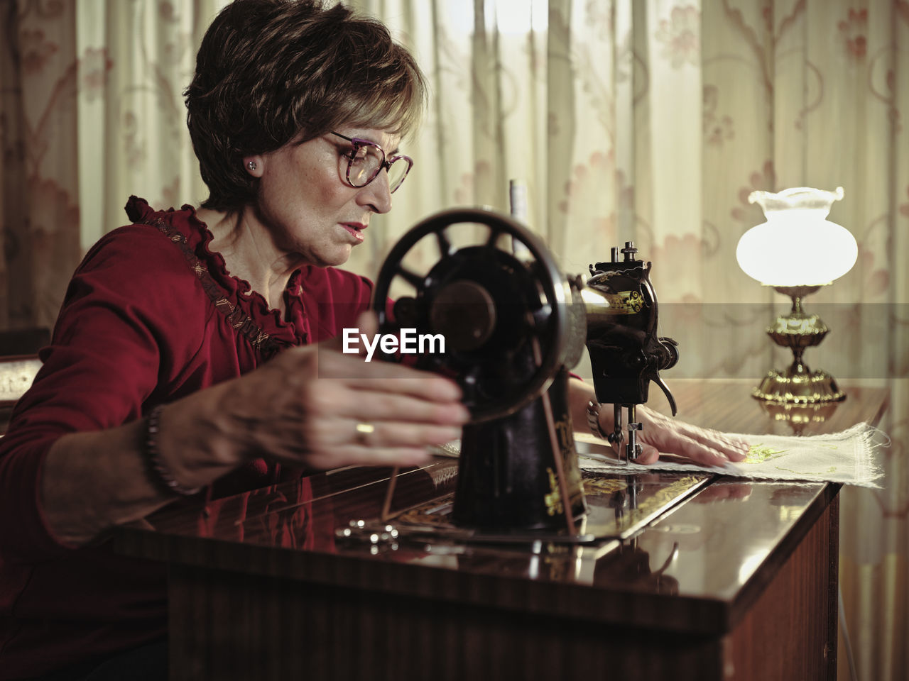 Senior lady in glasses using retro sewing machine to create linen napkin in cozy room at home