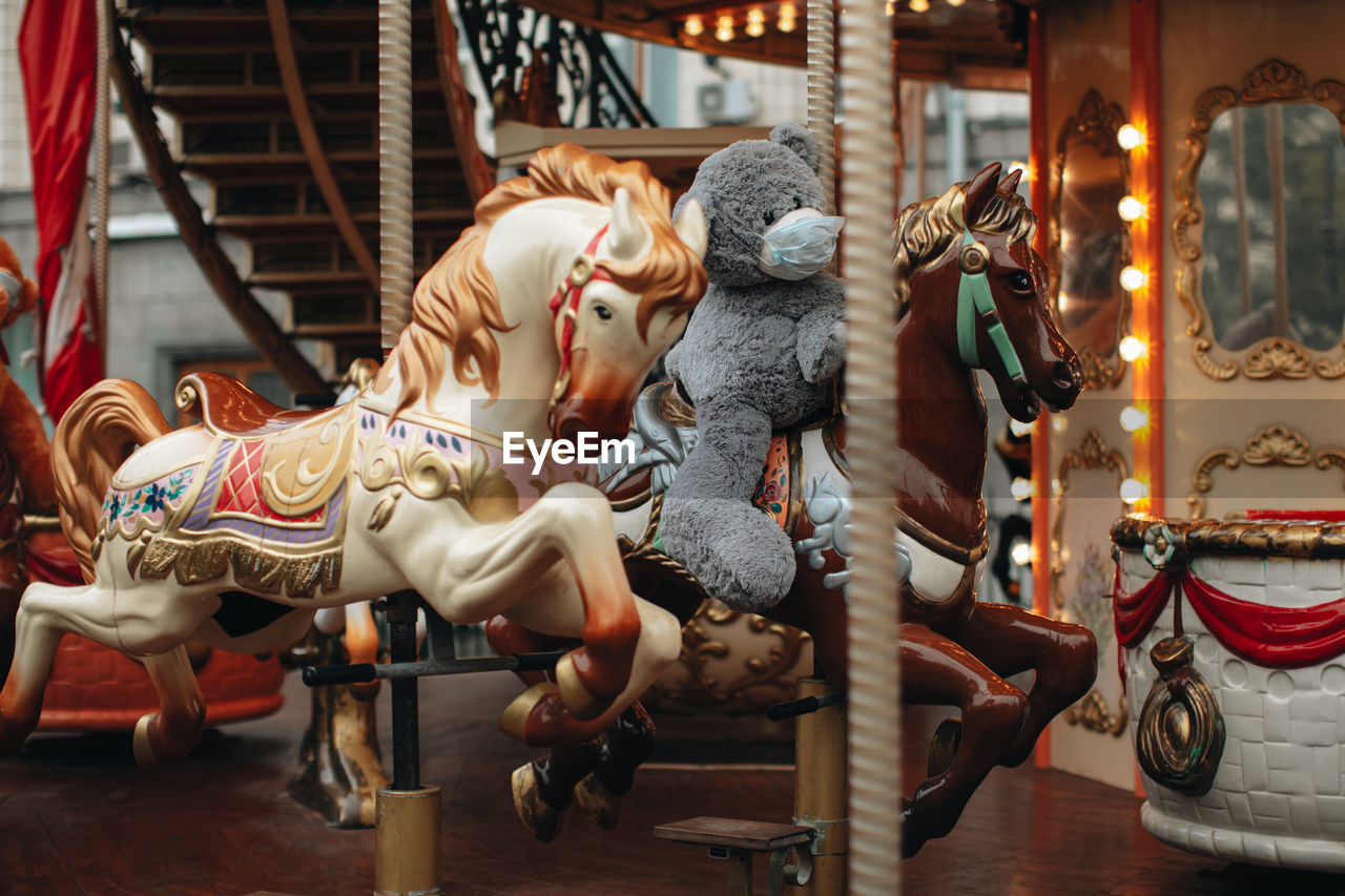 Children ride horses and teddy bear in protective mask on children's carousels in an amusement park.