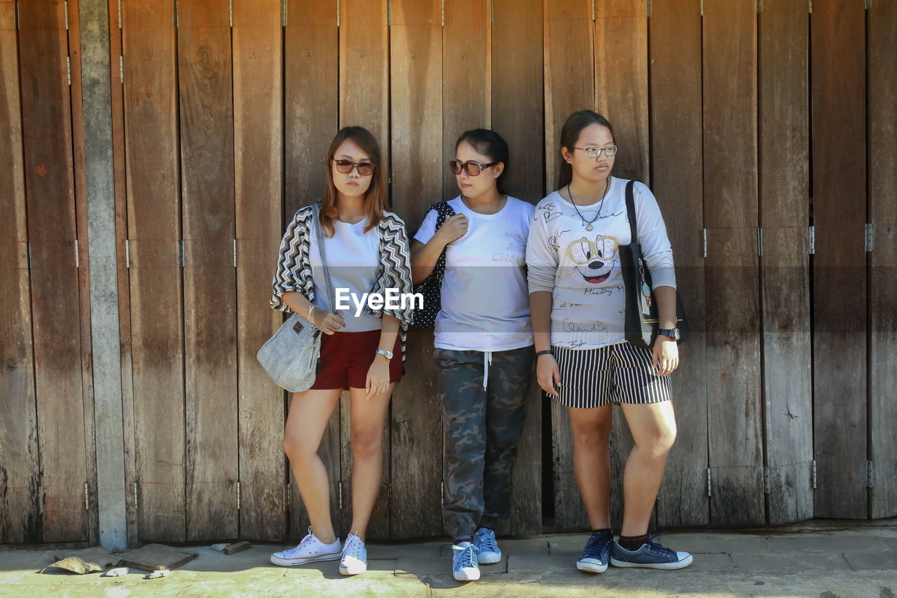 Full length of sisters standing with mother against wooden fence