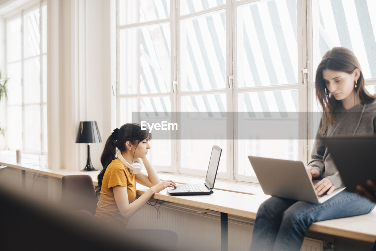 Female colleagues using laptops on desk against window at office