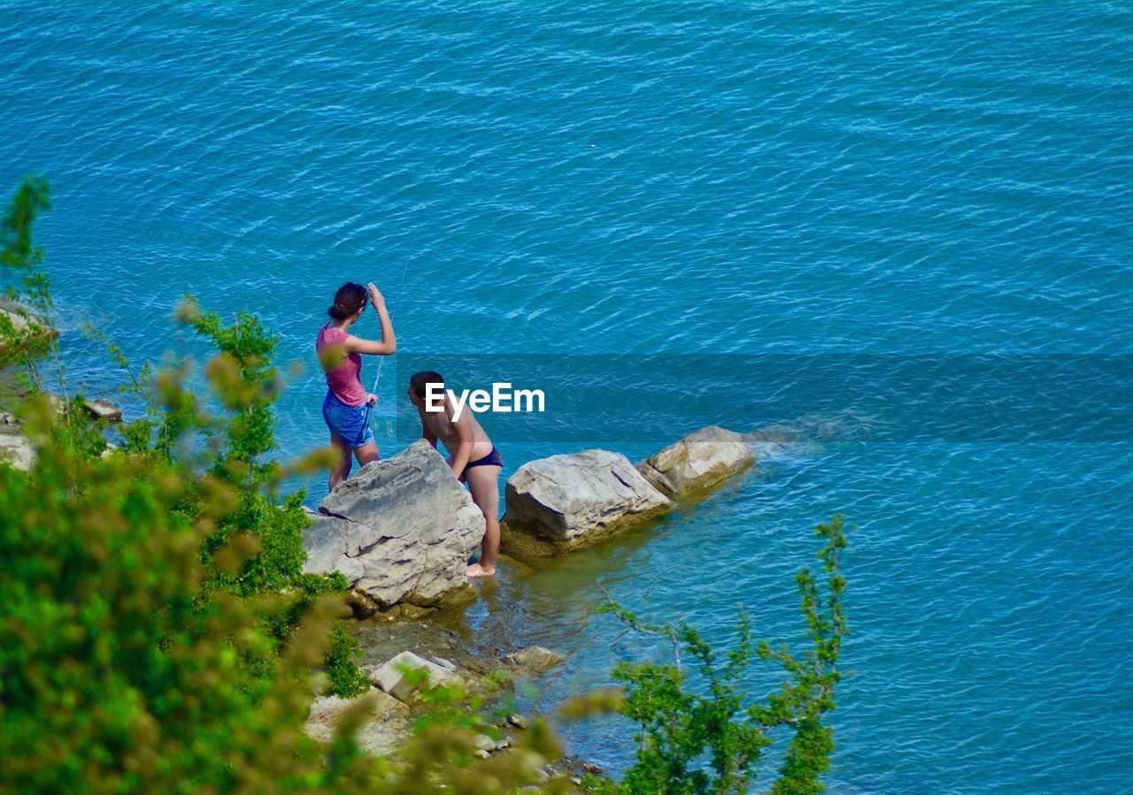 HIGH ANGLE VIEW OF FRIENDS RELAXING ON ROCK BY SEA