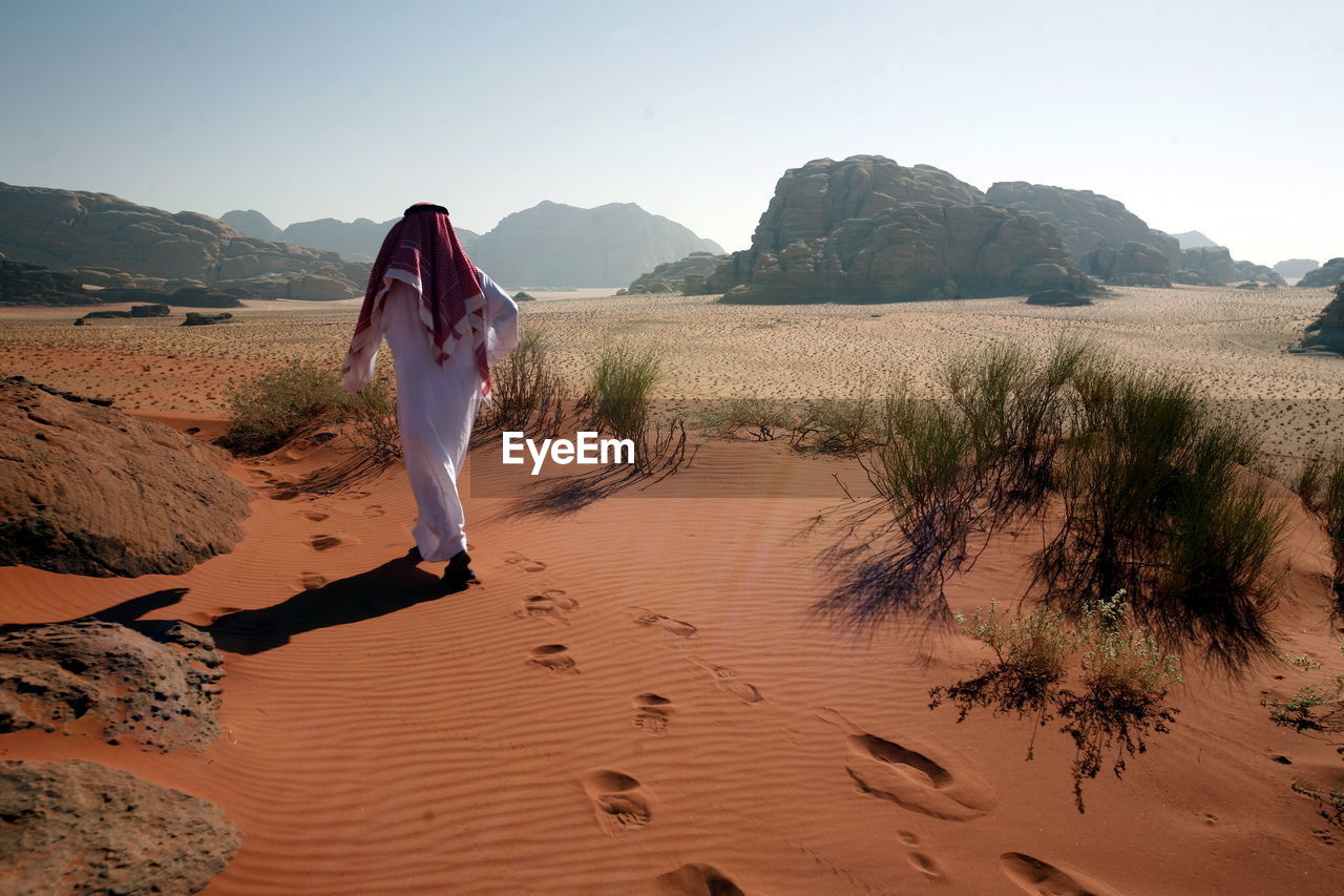 Rear view of man walking on sand dune against clear sky