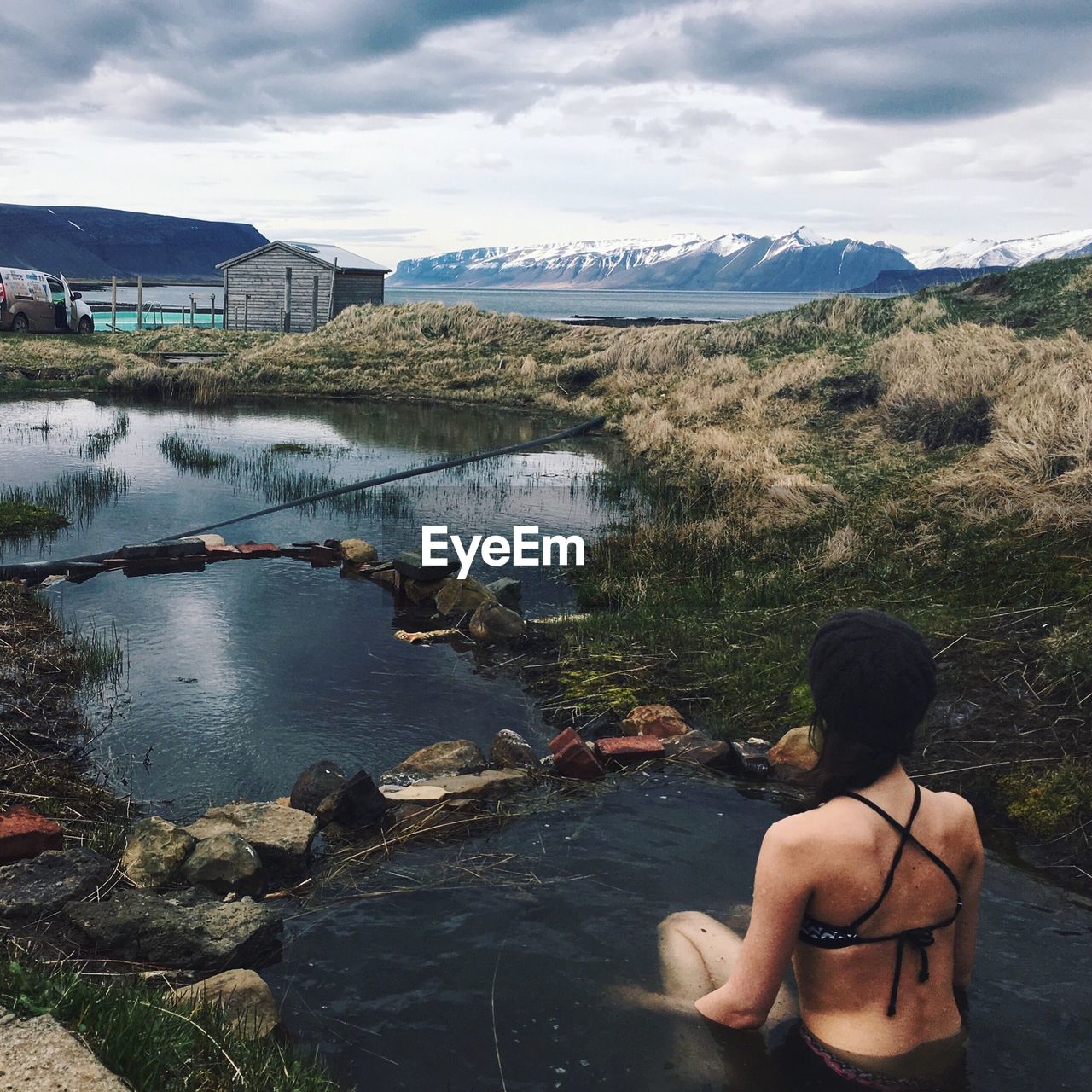 REAR VIEW OF MAN SITTING ON RIVER AGAINST SKY