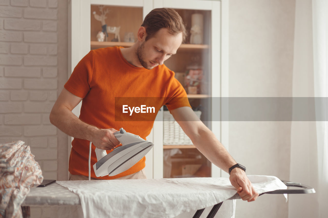 Young caucasian man ironing children's sheet on ironing board at home. men doing home chores